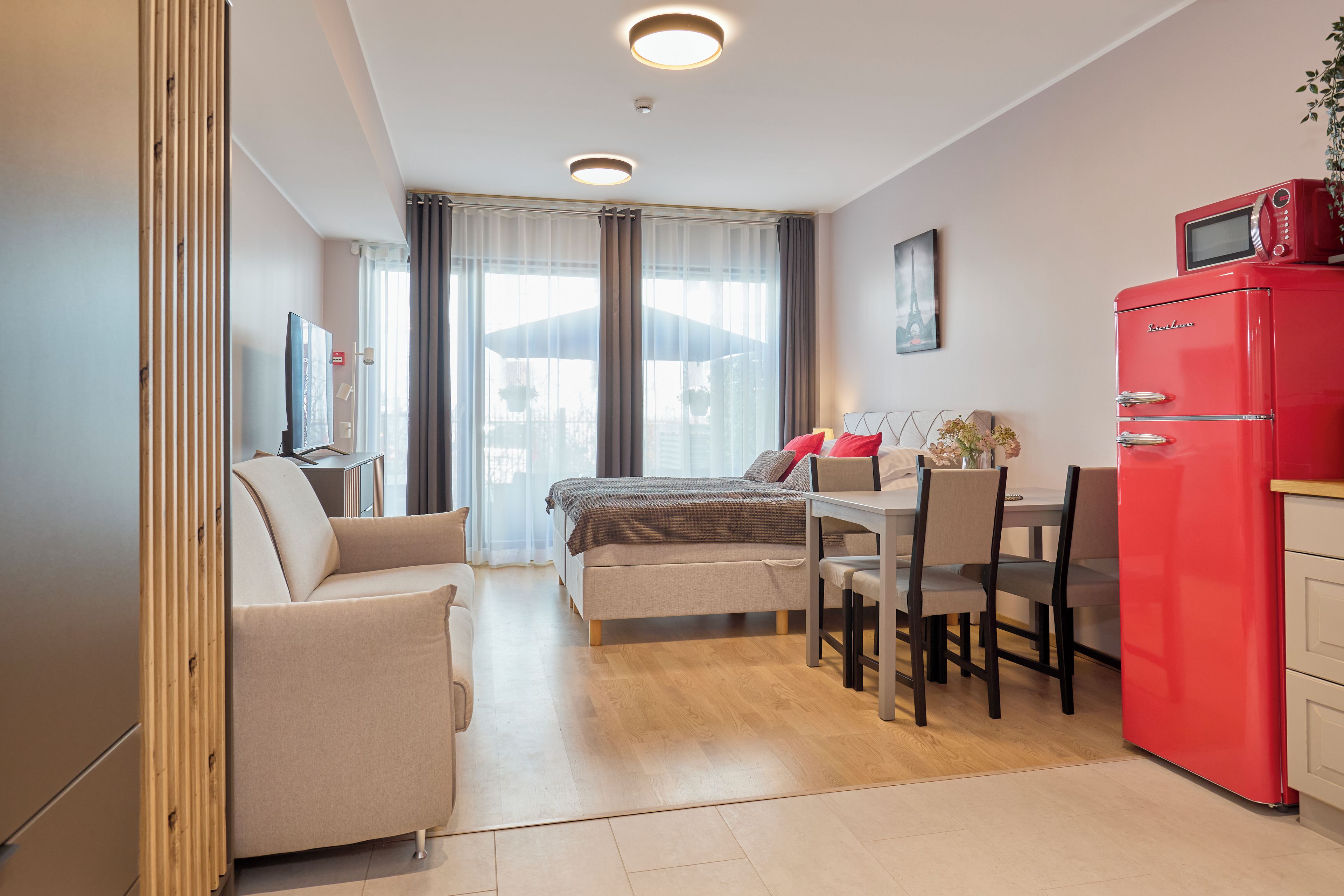 A compact hotel room with a bed, a desk, a chair, a red minibar, and a view through the window.
