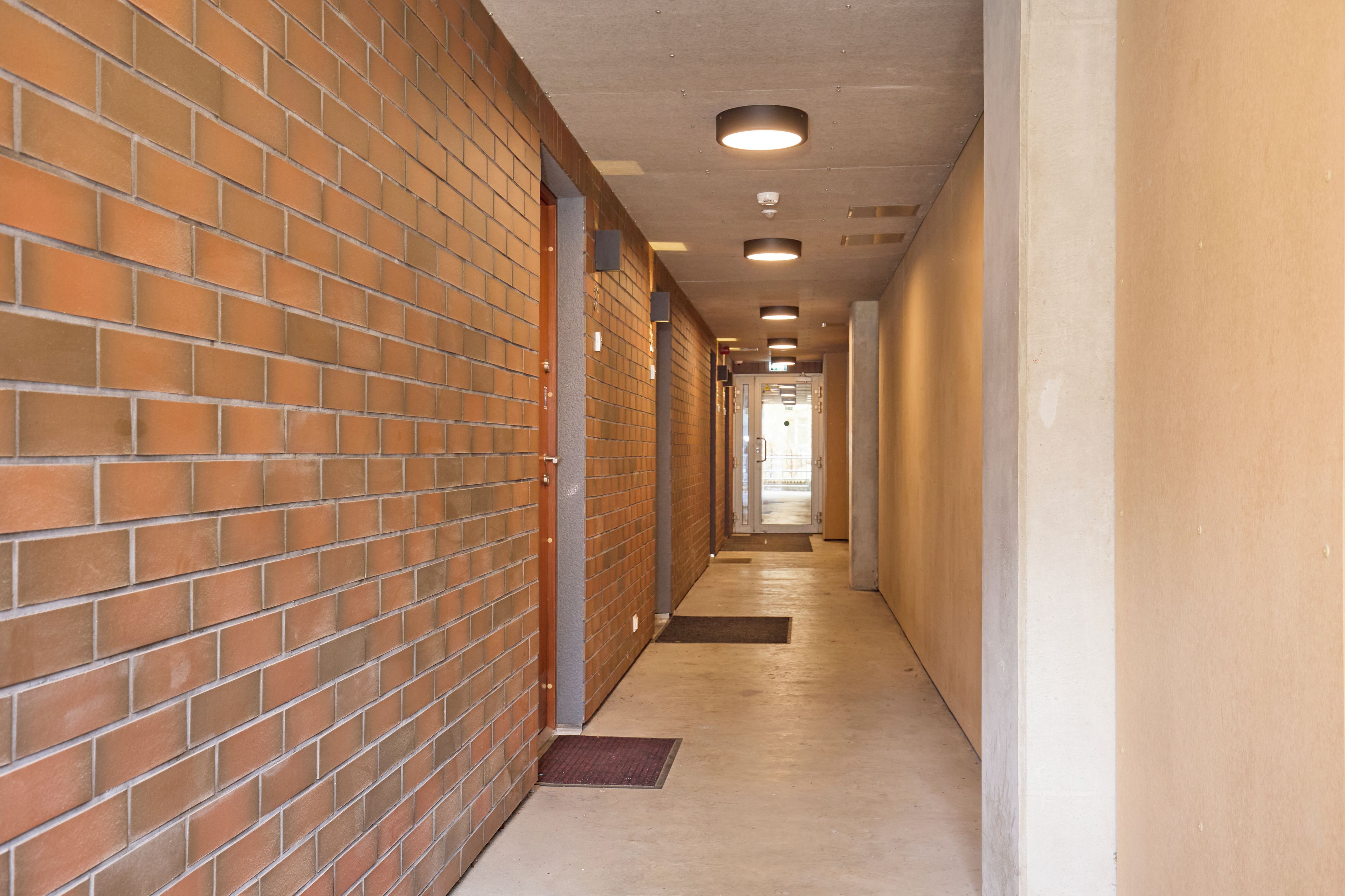 A corridor with brick walls and a tiled floor, leading to a door at the end.