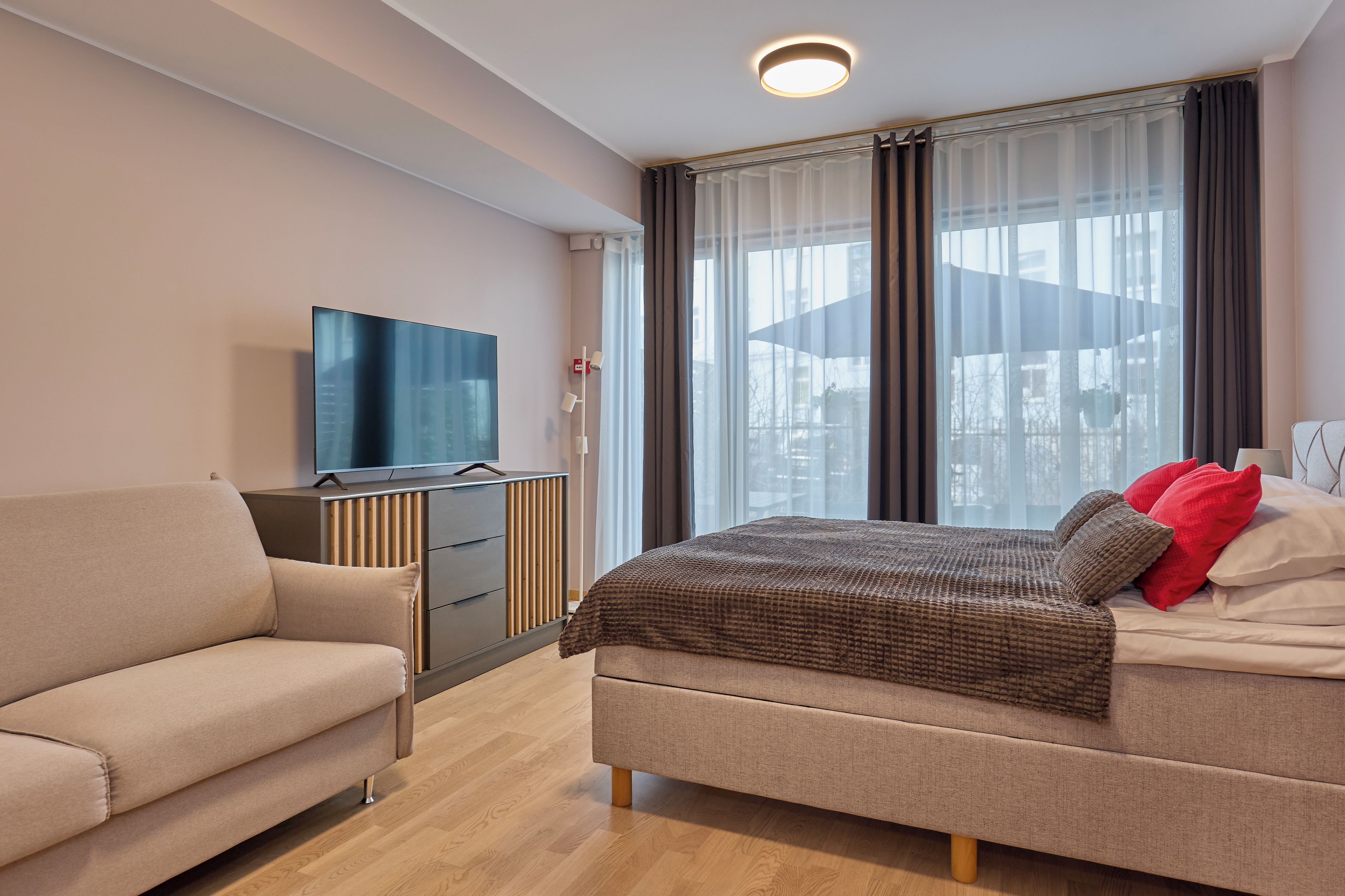 A modern hotel room with a bed, a sofa, a TV, and a window with curtains.