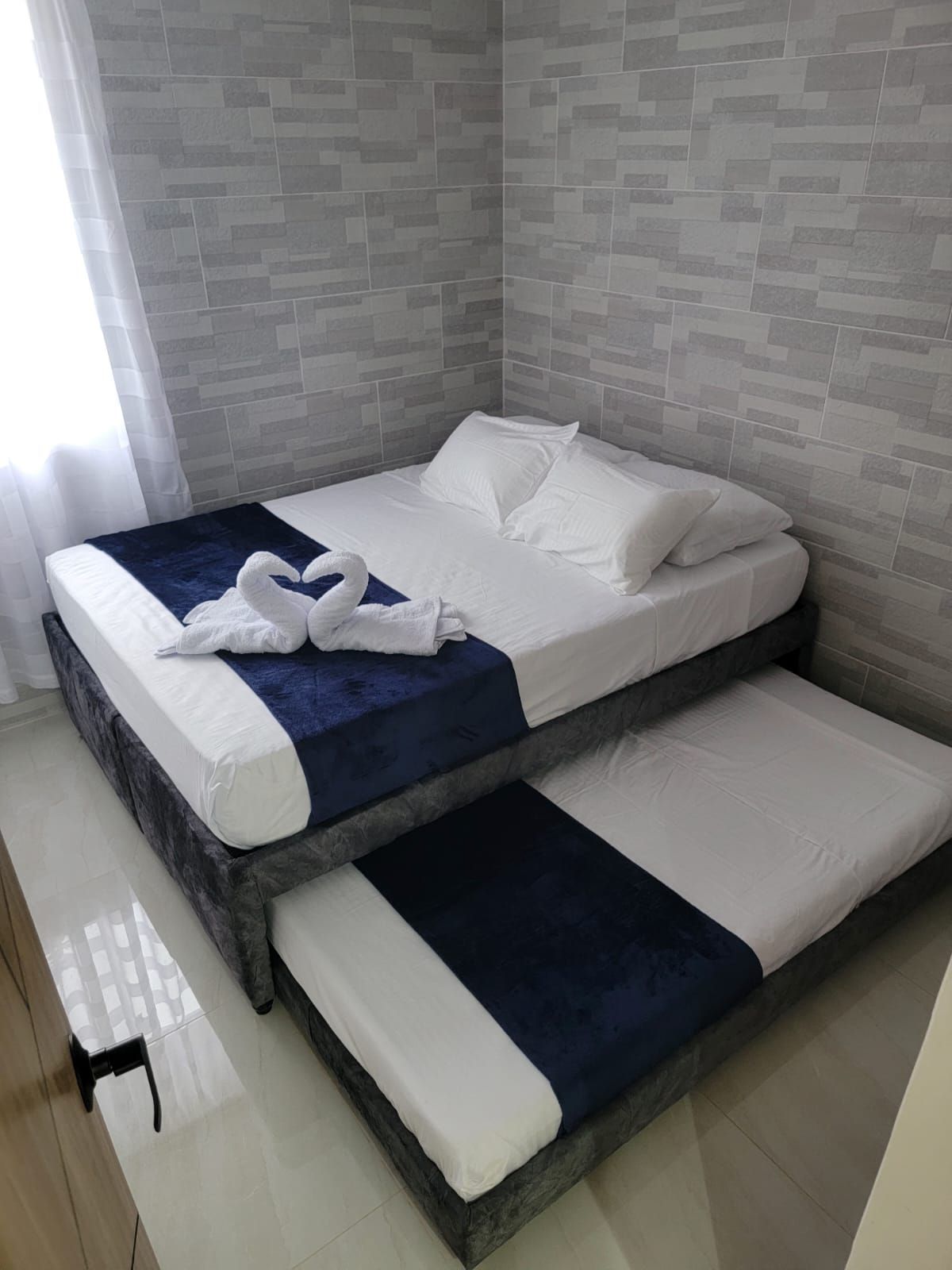 A double bed with white linens and a blue accent, positioned in a room with grey tiled walls and a wooden floor.