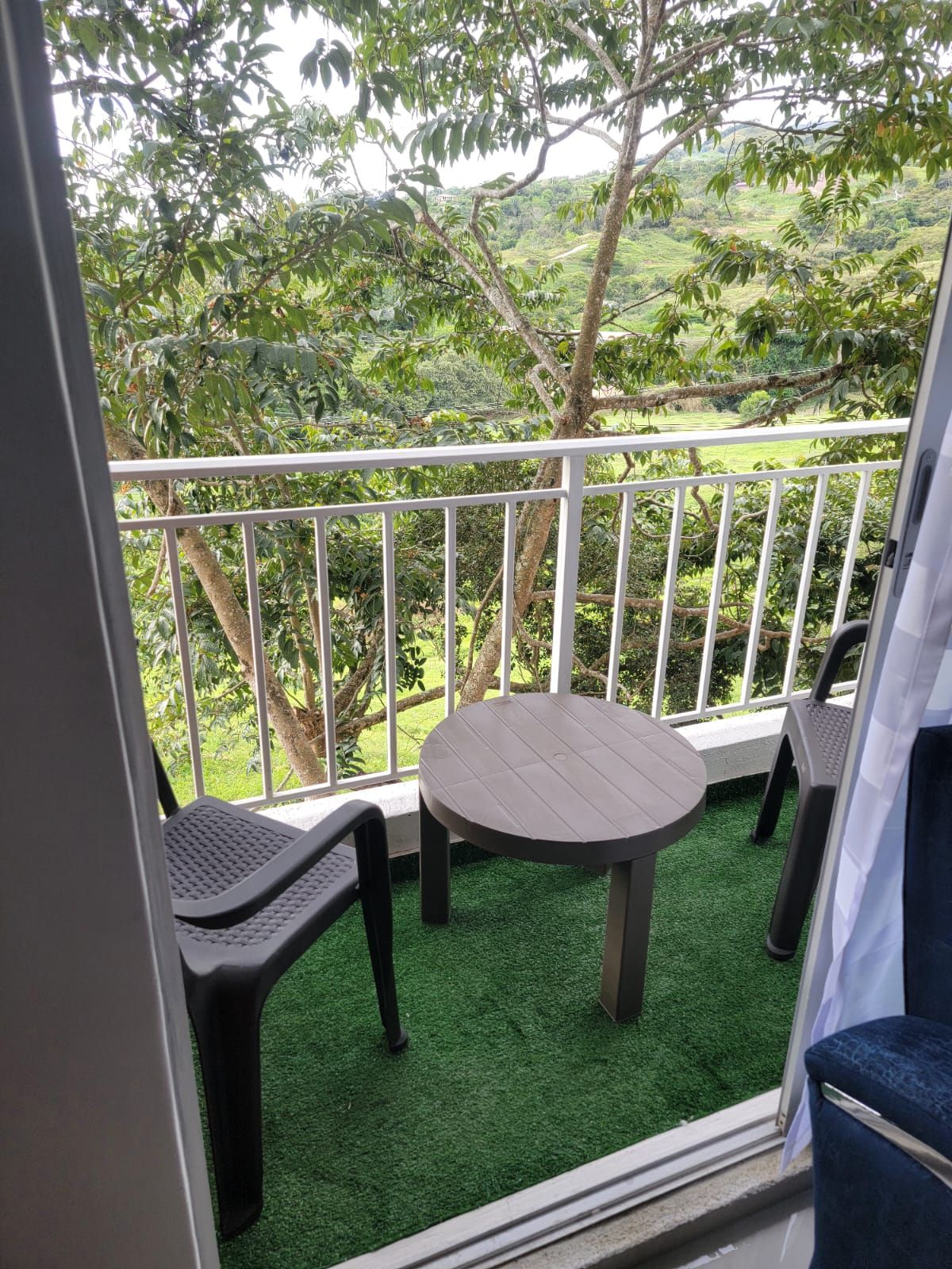 Ein Balkon mit rundem Tisch und zwei Stühlen, umgeben von einem weißen Geländer, mit Blick auf eine üppige grüne Landschaft.