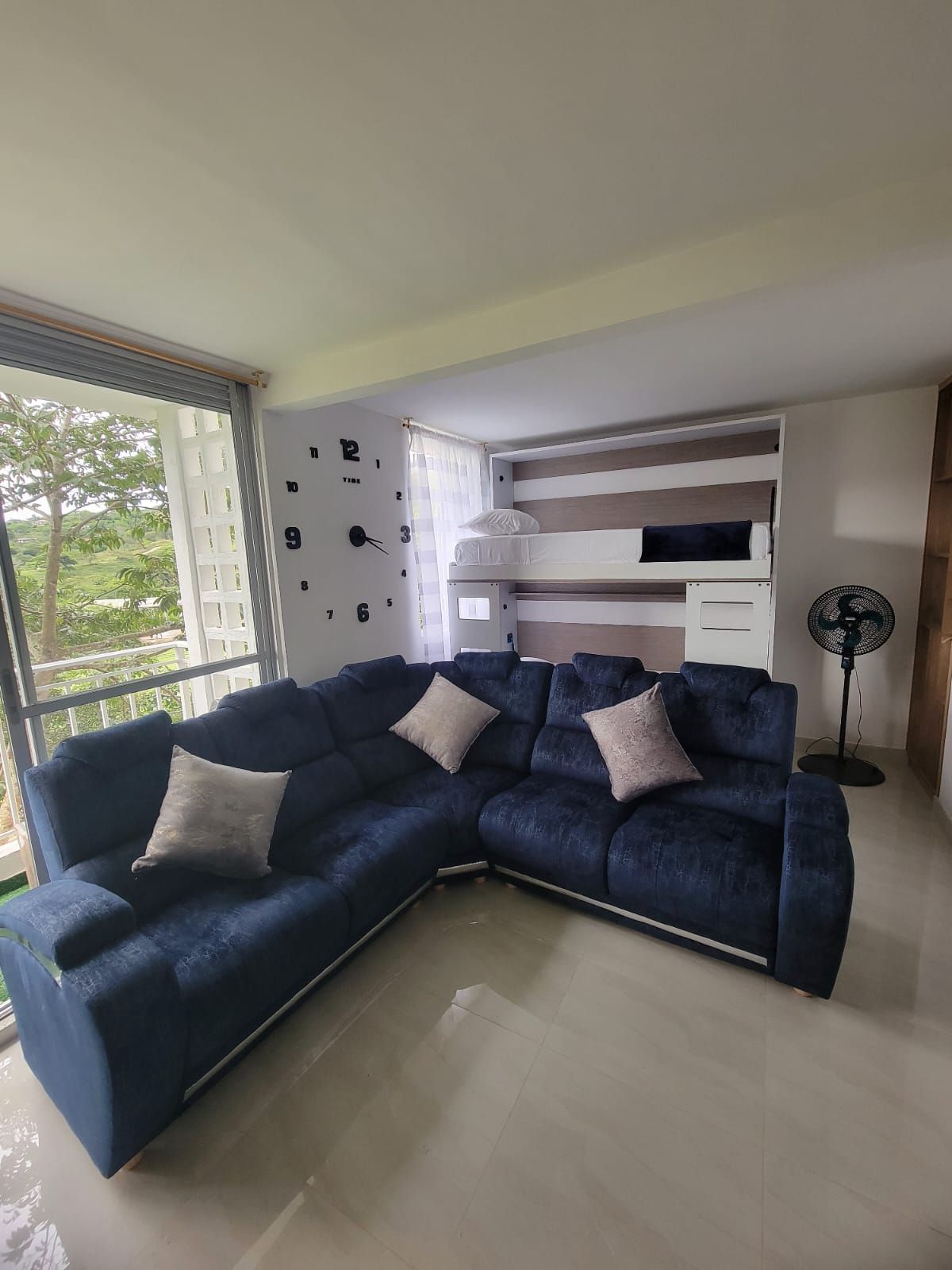 Ein modernes Wohnzimmer mit einem blauen Ecksofa, einer eingebauten Bank, einer Wanduhr und einem Standventilator.