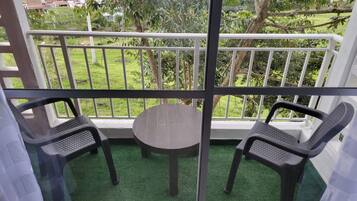 Un balcone con un tavolo e due sedie, delimitato da una ringhiera bianca, con vista su una zona verde con alberi e edifici.