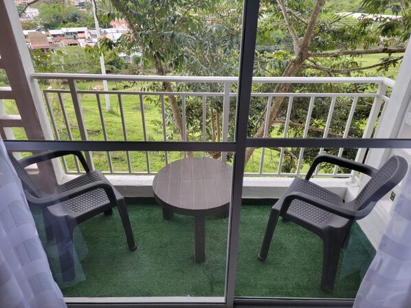 A balcony with a table and two chairs, surrounded by a white railing, overlooking a green area with trees and buildings.