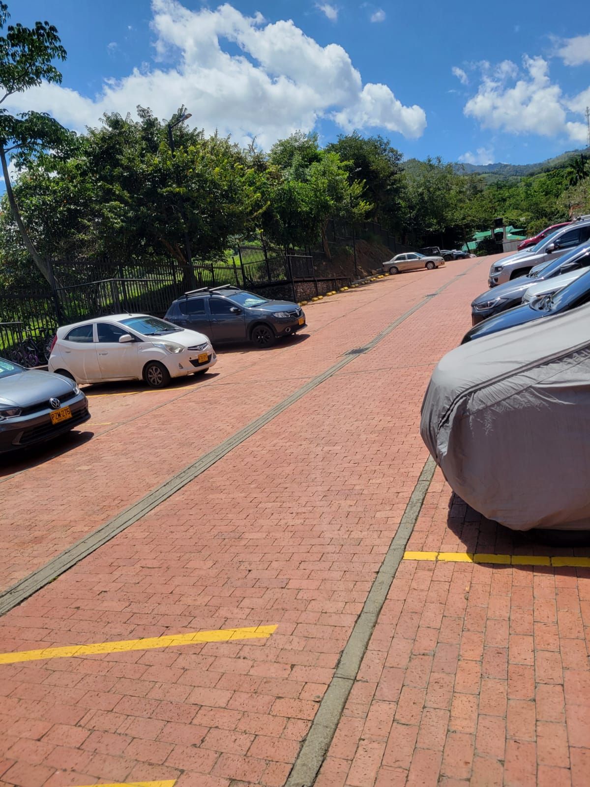 Un estacionamiento con varios autos estacionados sobre una superficie pavimentada, rodeado de árboles y una cerca.