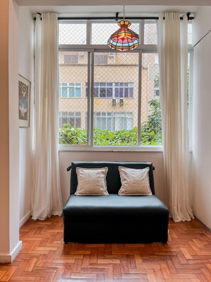 A room with a black sofa, two patterned cushions, a stained glass window, and a view of buildings and greenery outside. - Copacabana Refuge Minutes From the Sea Jc602 (Rio de Janeiro)