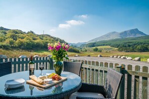 A table with a bottle, glass, cheese, fruit, and flowers set against a scenic mountain backdrop.