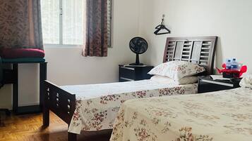Quarto com duas camas, uma cadeira, mesa de cabeceira, ventilador e janela com cortinas.