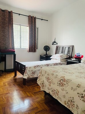 Quarto com duas camas, uma cadeira, mesa de cabeceira, ventilador e janela com cortinas.