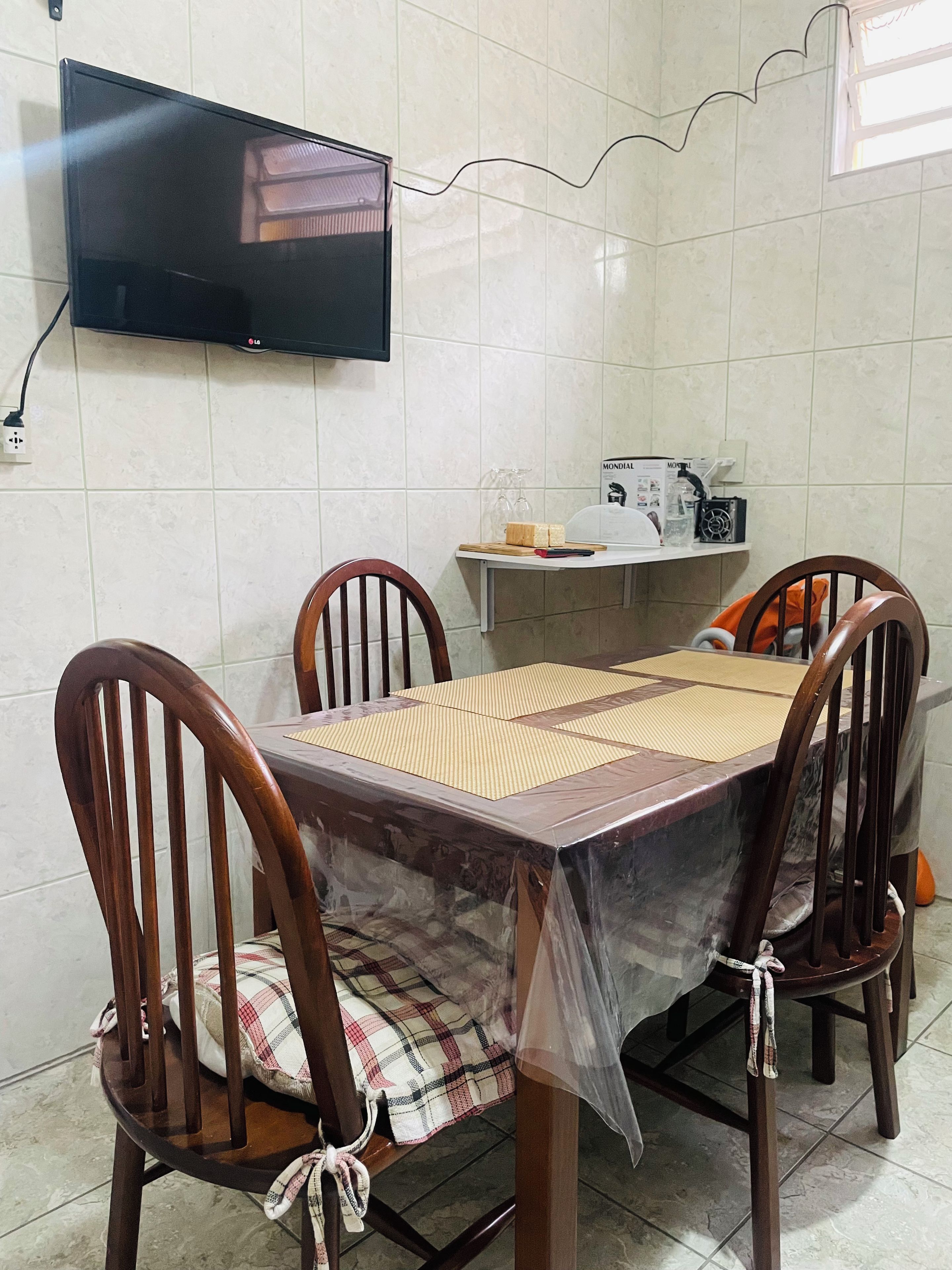 Uma área de refeições com uma mesa coberta por plástico, quatro cadeiras de madeira, uma televisão na parede e uma prateleira com utensílios de cozinha.