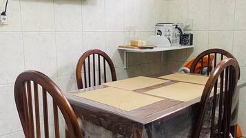 Uma área de refeições com uma mesa coberta por plástico, quatro cadeiras de madeira, uma televisão na parede e uma prateleira com utensílios de cozinha.