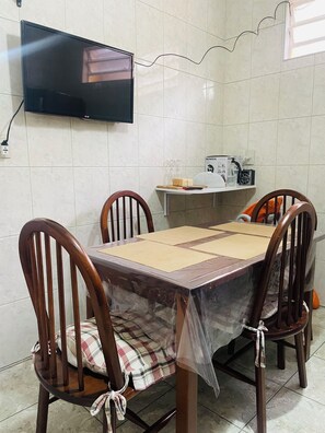 Uma área de refeições com uma mesa coberta por plástico, quatro cadeiras de madeira, uma televisão na parede e uma prateleira com utensílios de cozinha.