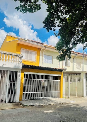 Edifício residencial com fachada amarela, entrada com portão e varanda visível.
