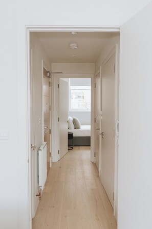 A narrow hallway leading to a bedroom with a bed, a radiator, and a window with a view of buildings. - Premium London Stay – Luxury 1-Bed Apartment (London)