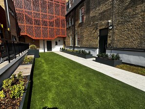 A courtyard with artificial grass, a brick building, and a modern red structure. - Premium London Stay – Luxury 1-Bed Apartment (London)
