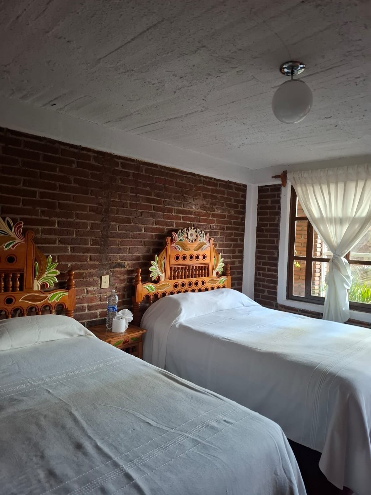 A room with two beds, a brick wall, a wooden headboard, a window with curtains, and a ceiling fan.