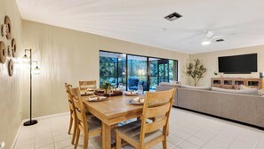 A dining area with a wooden table and chairs, a sofa, and a television. - Coralvine by Avantstay Screened Heated Pool + Lanai & Grill Near Beach + Mercato (Naples)