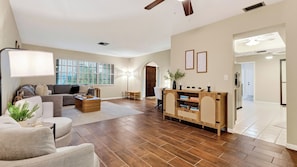 A spacious living room with hardwood flooring, a sofa, a coffee table, and a wooden cabinet with books. - Coralvine by Avantstay Screened Heated Pool + Lanai & Grill Near Beach + Mercato (Naples)