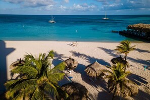 Een strand met helder turquoise water, een stel dat over het zand loopt, en rieten parasols.