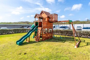 A wooden play structure with a slide and climbing area, surrounded by a grassy field and parked cars. - PC404 - Pool View - 1 BR Perfect  for a couple - Close to Palm Beach - 1BR (Palm Beach)
