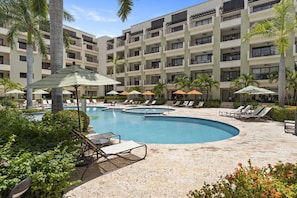Ein Hotelpoolbereich mit mehreren Liegestühlen und Sonnenschirmen, umgeben von Palmen und einem mehrstöckigen Gebäude.