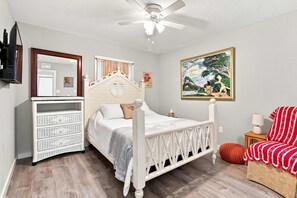 A bedroom with a bed, dresser, ceiling fan, artwork, and a chair.