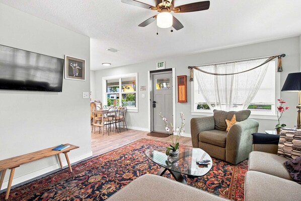 A living room with a ceiling fan, a dining area with a table and chairs, and a view of the outside through a window.