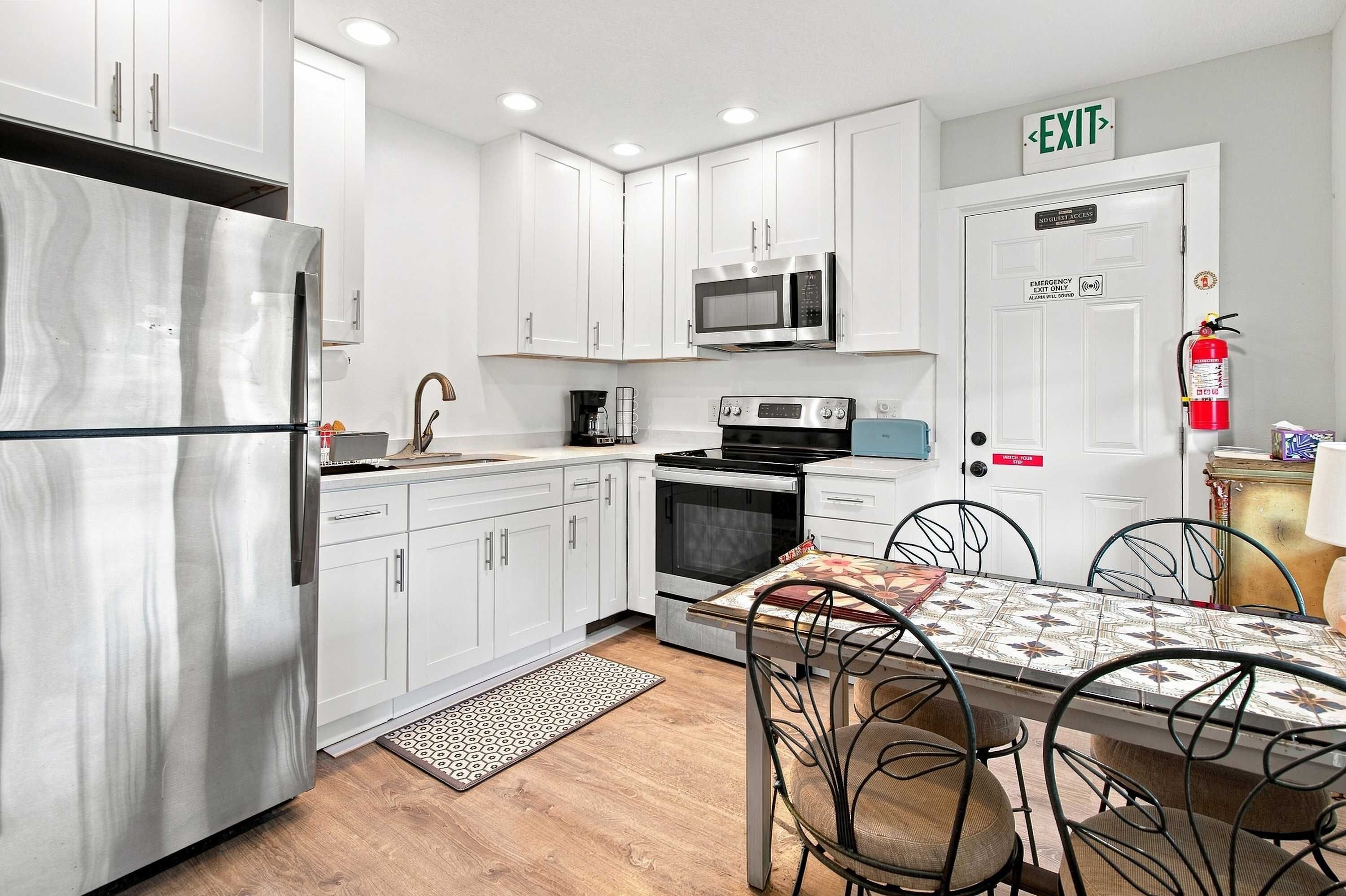 A compact kitchen with white cabinets, a stainless steel refrigerator, a microwave, an oven, and a coffee maker. There's a dining table with chairs and a patterned table runner.