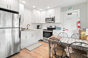 A compact kitchen with white cabinets, a stainless steel refrigerator, a microwave, an oven, and a coffee maker. There's a dining table with chairs and a patterned table runner.