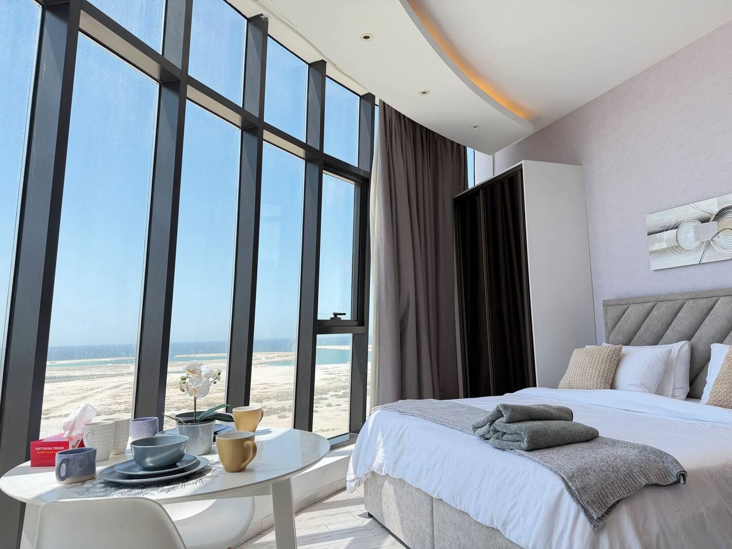 A hotel room with a large window, a bed, a bedside table, and a view of the beach.