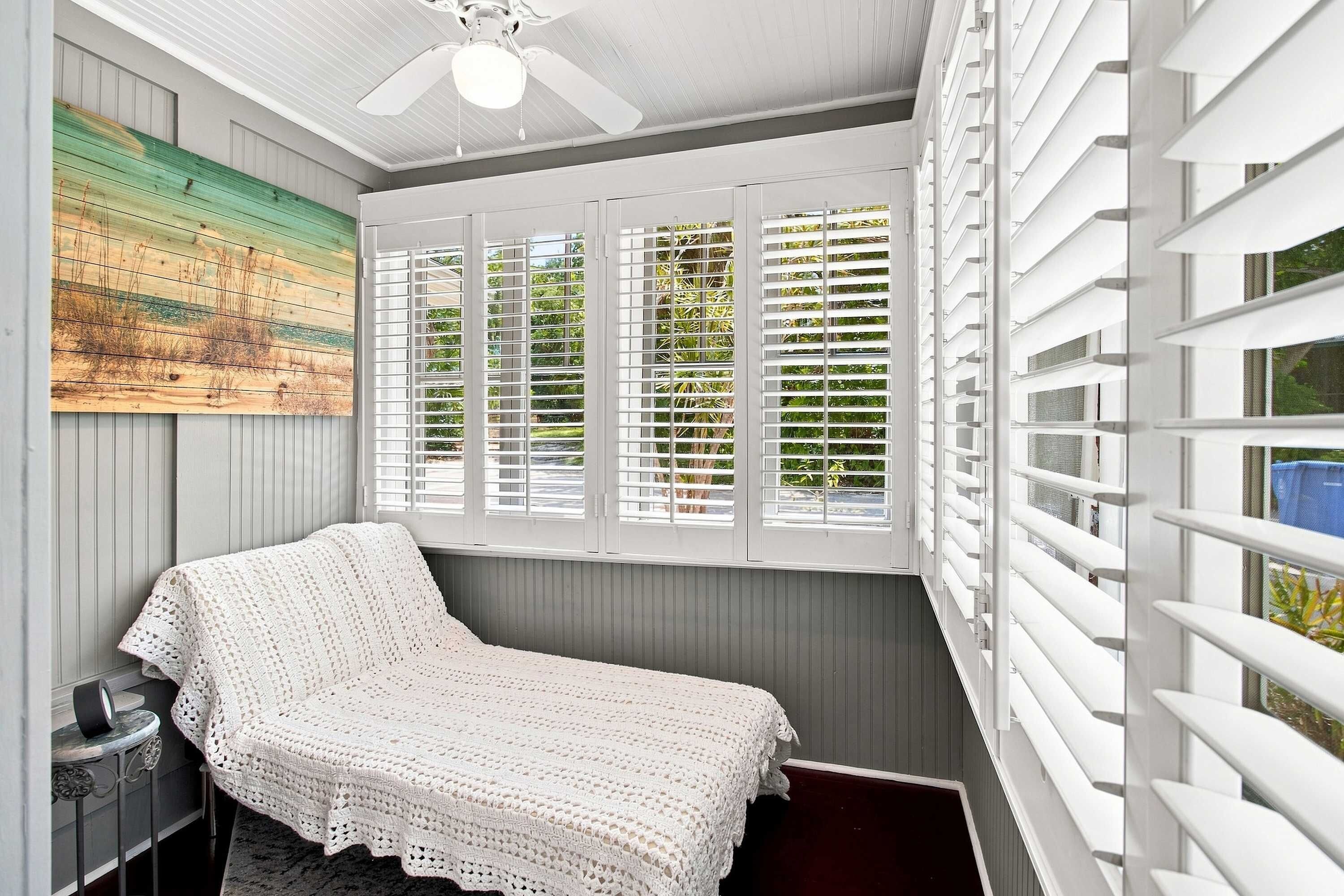 A room with a bed, a ceiling fan, a window with blinds, and a painting on the wall.