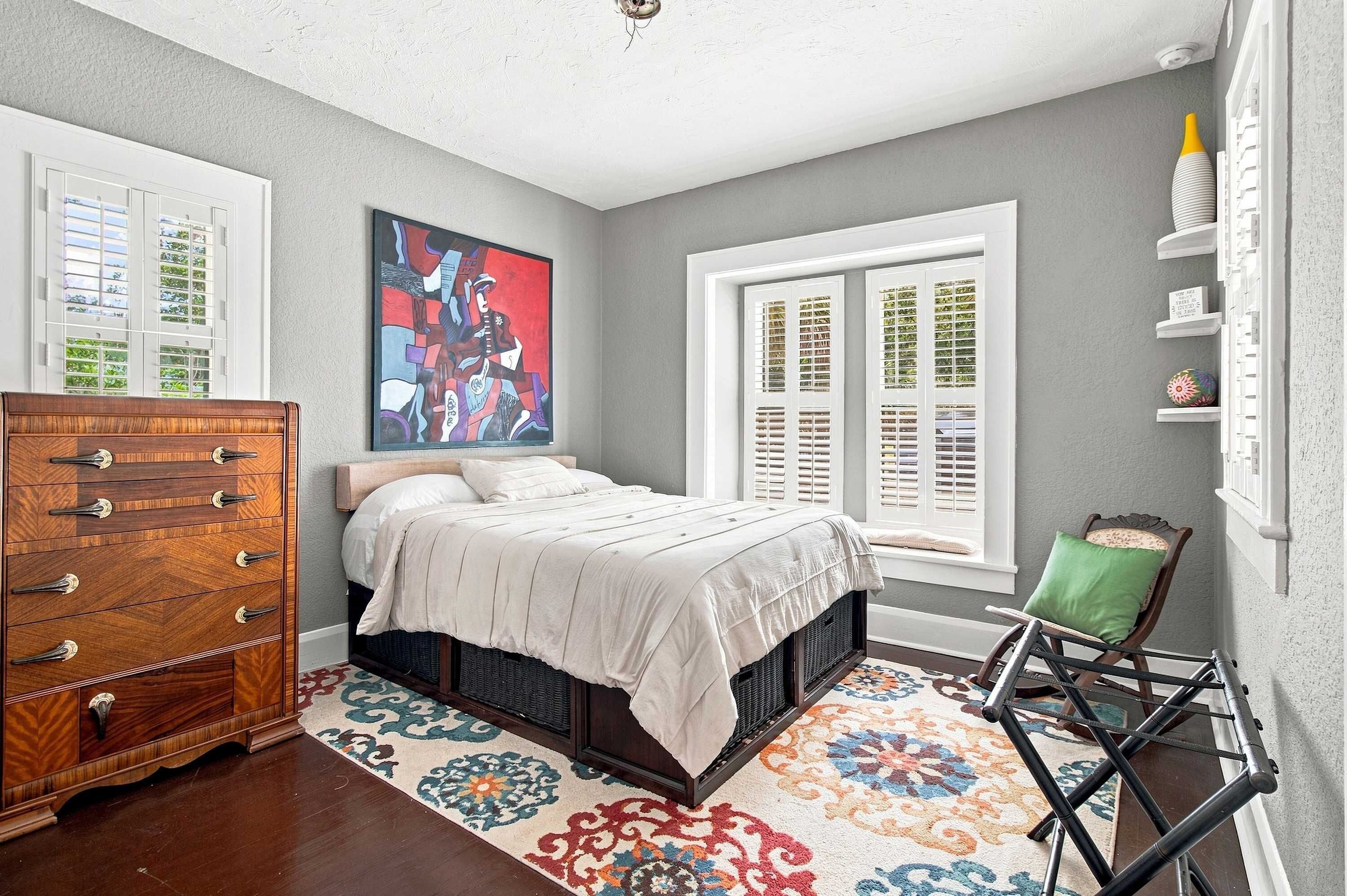 A bedroom with a bed, a dresser, a colorful rug, a window with shutters, and a painting on the wall.