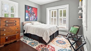 Um quarto com cama, penteadeira, tapete colorido, janela com persianas e um quadro na parede.