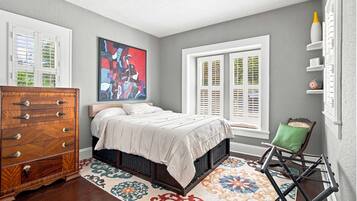 A bedroom with a bed, a dresser, a colorful rug, a window with shutters, and a painting on the wall.