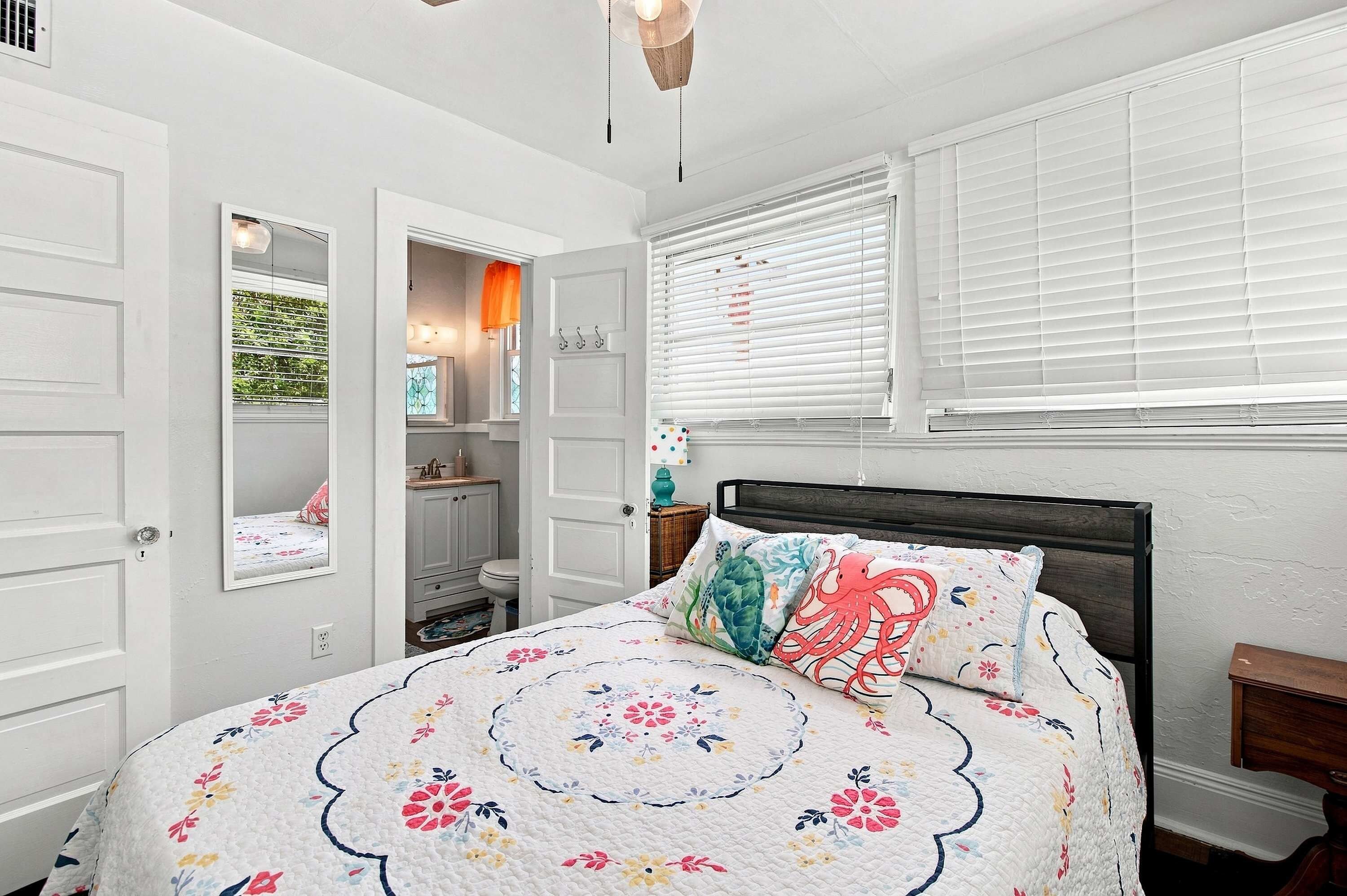 A bedroom with a bed, a nightstand, a window with blinds, and a door leading to a bathroom.