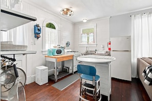 A compact kitchen with white cabinetry, a microwave, a sink, and a small dining area with a blue chair. - The Koi House Cottage (Bradenton)