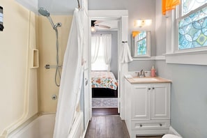 A narrow bathroom with a shower, a bed, a sink, and a mirror. - The Koi House Cottage (Bradenton)