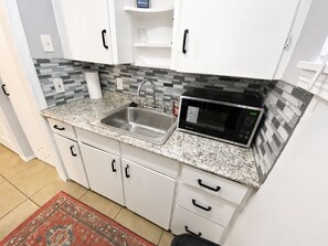 A kitchen with white cabinets, a granite countertop, a stainless steel sink, a black microwave, and a patterned rug. - Vintage Charm Meets Cozy Comfort in this Character-Filled Old East Dallas Studio (Dallas)
