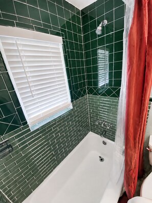 A bathroom with a white bathtub, green mosaic tiles, a white window with blinds, and a red shower curtain. - Vintage Charm Meets Cozy Comfort in this Character-Filled Old East Dallas Studio (Dallas)