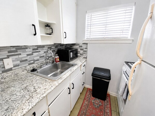 A compact kitchen with a granite countertop, stainless steel sink, white cabinets, a black trash bin, a microwave, and a window with blinds. - Vintage Charm Meets Cozy Comfort in this Character-Filled Old East Dallas Studio (Dallas)