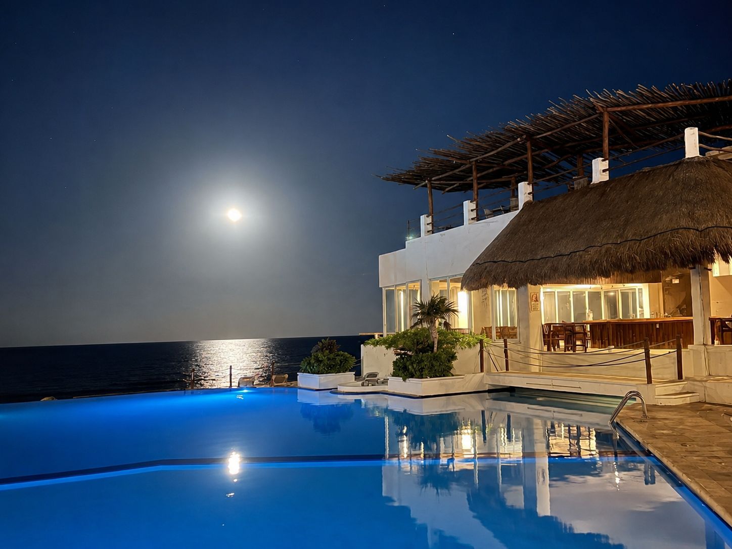 A nighttime pool area with a clear view of the ocean and a full moon.
