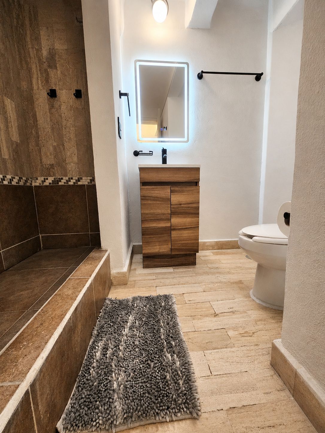 Ein Badezimmer mit einem Holzkosmetikschrank, einem beleuchteten Spiegel, einer Toilette und einer Duschkabine.