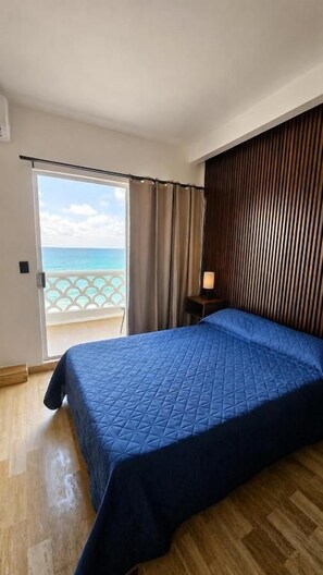 Quarto com cama, mesa de cabeceira, luminária, porta de vidro corrediça para uma varanda e vista para o oceano.