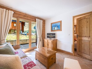 Een gezellige woonkamer met een houten salontafel, een bank met opvallende kussens, een televisie op een houten kast en een groot raam met gordijnen.