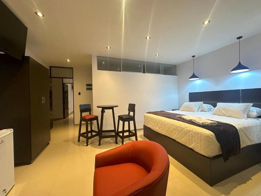 A modern hotel room with a large bed, a red chair, a small table, and a minibar.