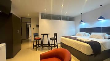 A modern hotel room with a large bed, a red chair, a small table, and a minibar.