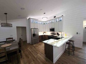 A modern kitchen with white cabinetry, a central island, and a dining area with wooden chairs. - 4 BR/3 Bath Tuscaloosa Retreat With Pool, Hot Tub and Putting Green (Northport)