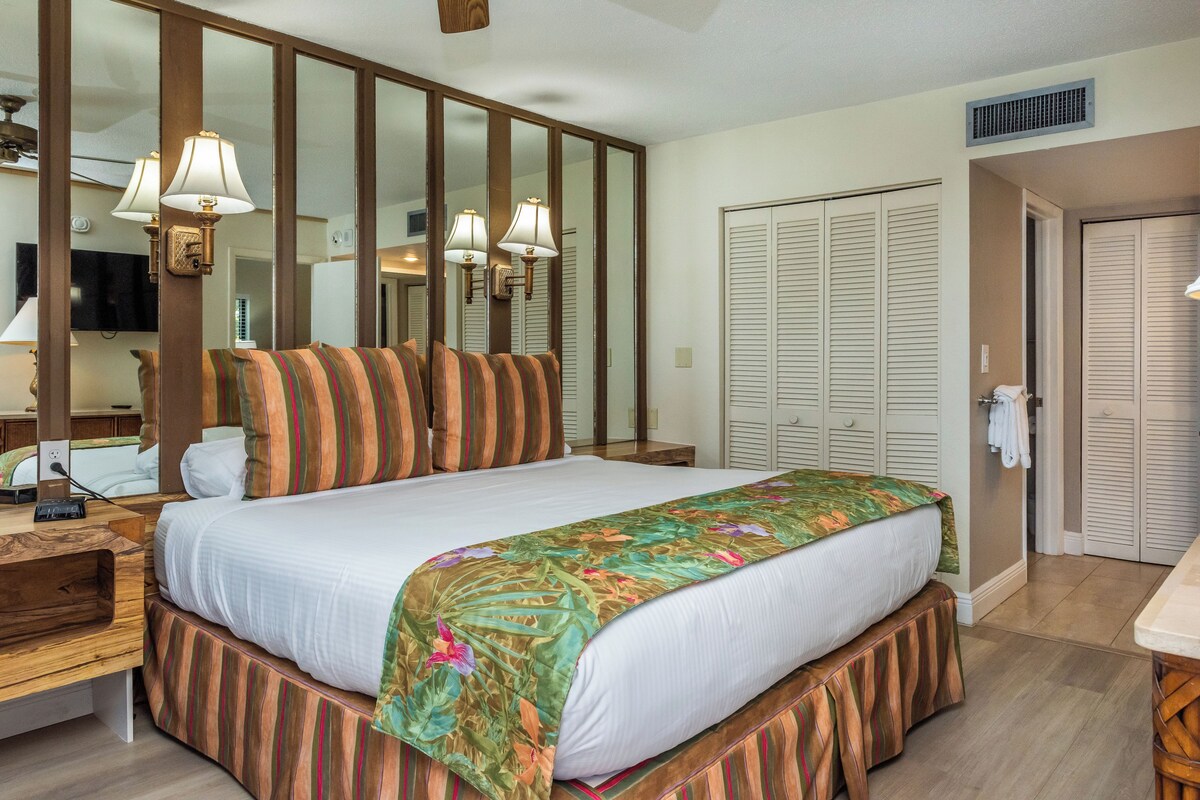 A hotel room with a large bed, a wooden nightstand, a desk with a telephone, and a wardrobe with louvered doors.