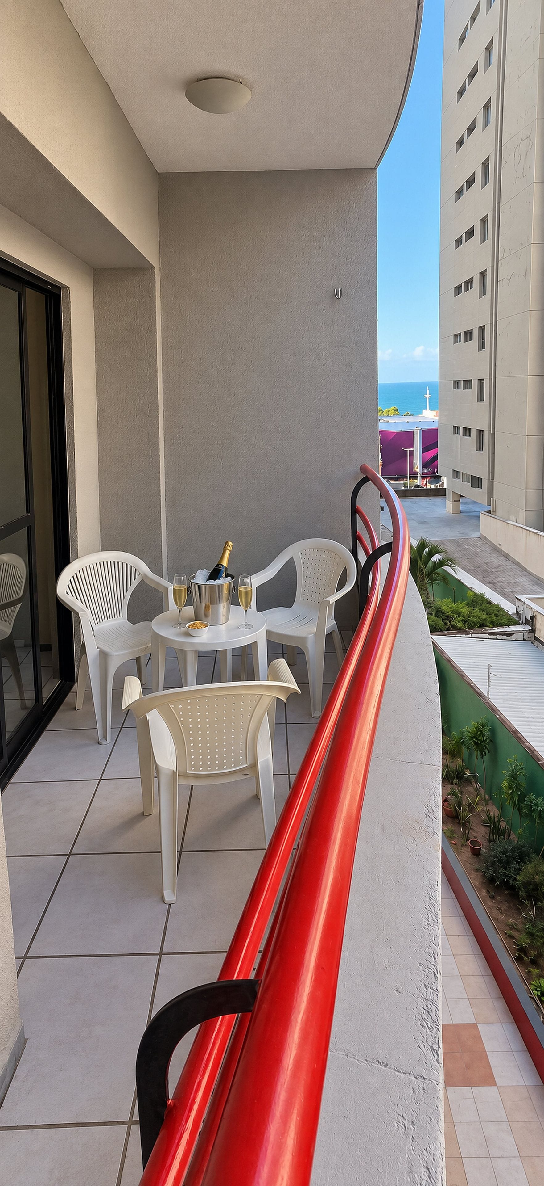An outdoor seating area with white plastic chairs and a table, a red metal slide, and a view of the sea and buildings in the background.