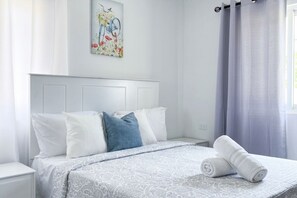 Ein ordentlich bezogenes Bett mit einem gemusterten Überwurf, weißen und blauen Kissen und aufgerollten weißen Handtüchern daneben.